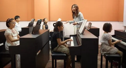 A group piano class for children, a teacher guiding several young students at keyboards